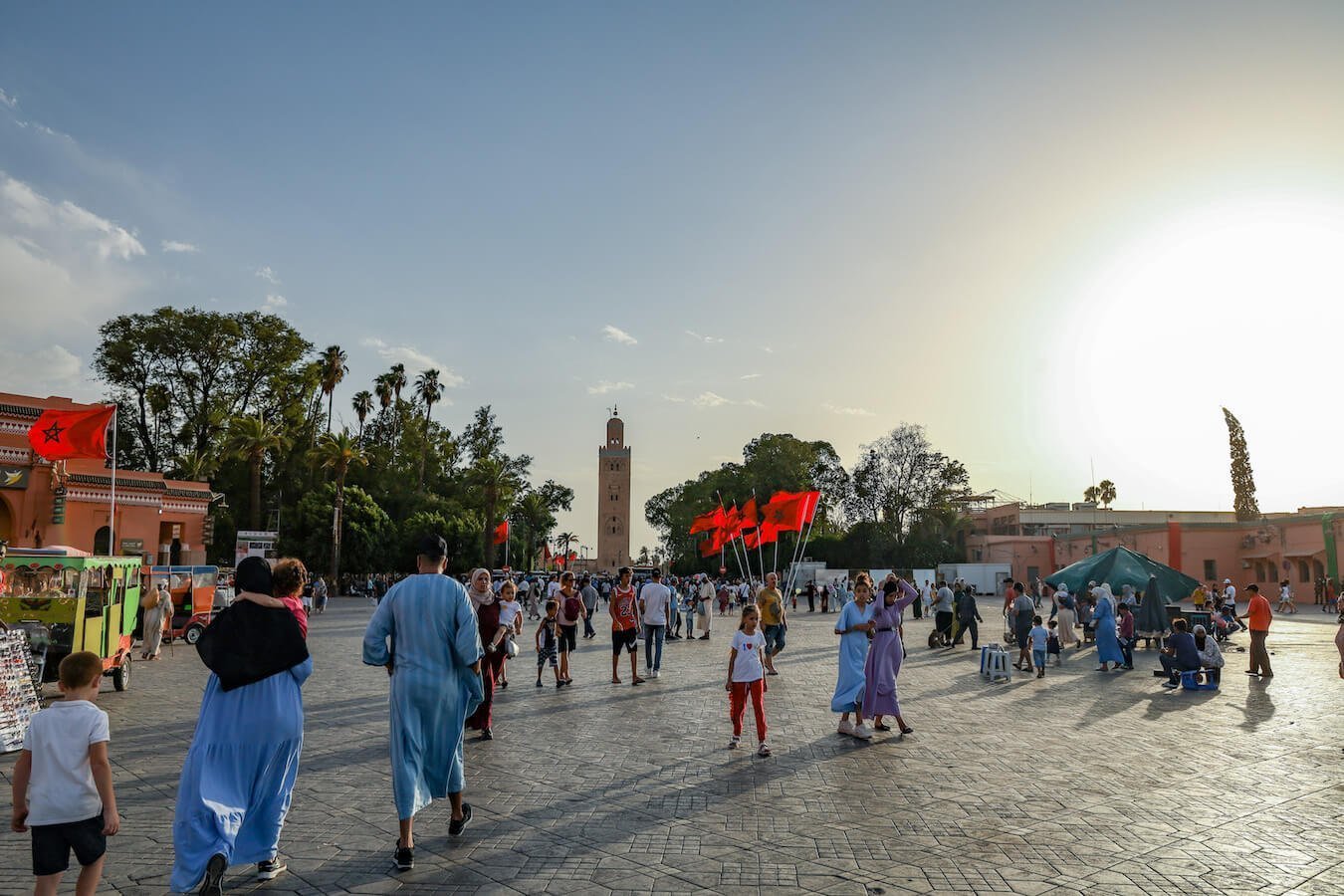 People walk around the Djemaa el-fna square in Marrakech with the Morocco flag flying and Koutoubia Mosque in the background