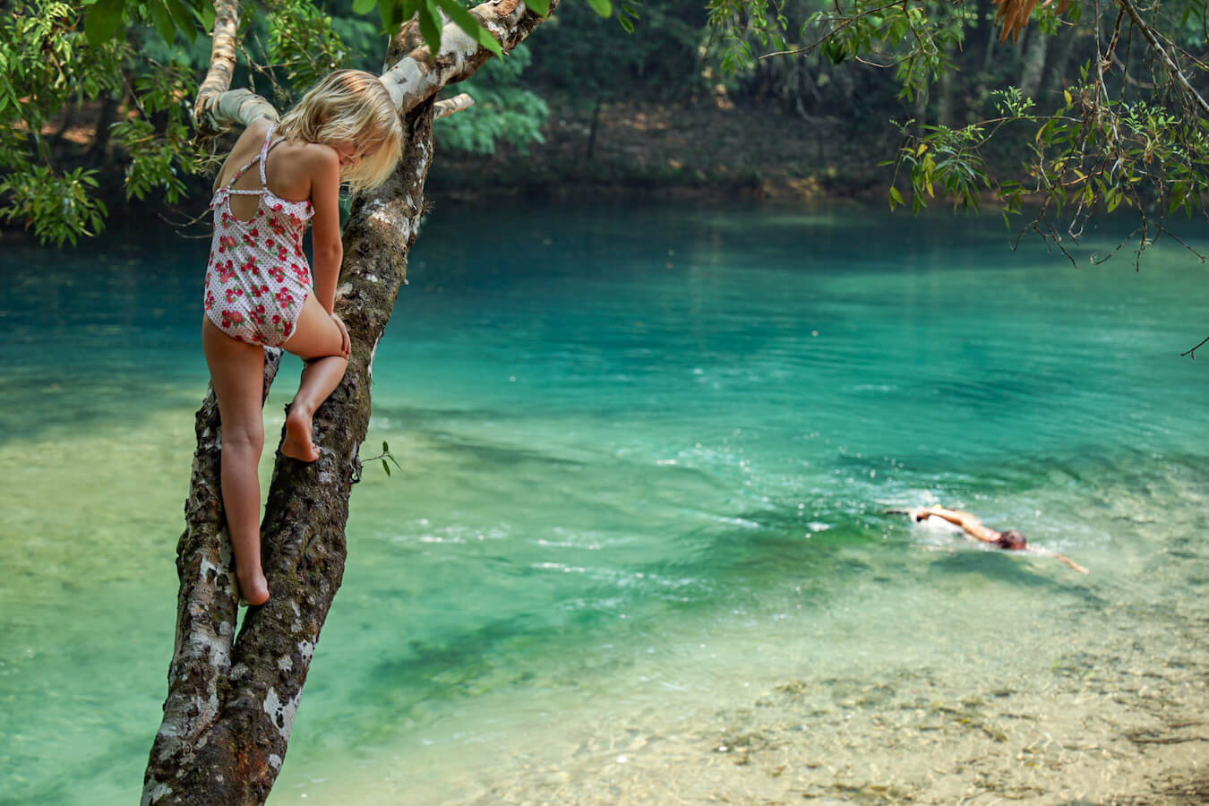 A young girl climbs a tree at Auga Azul in Palenque, while a man swims in the fresh water pool below.