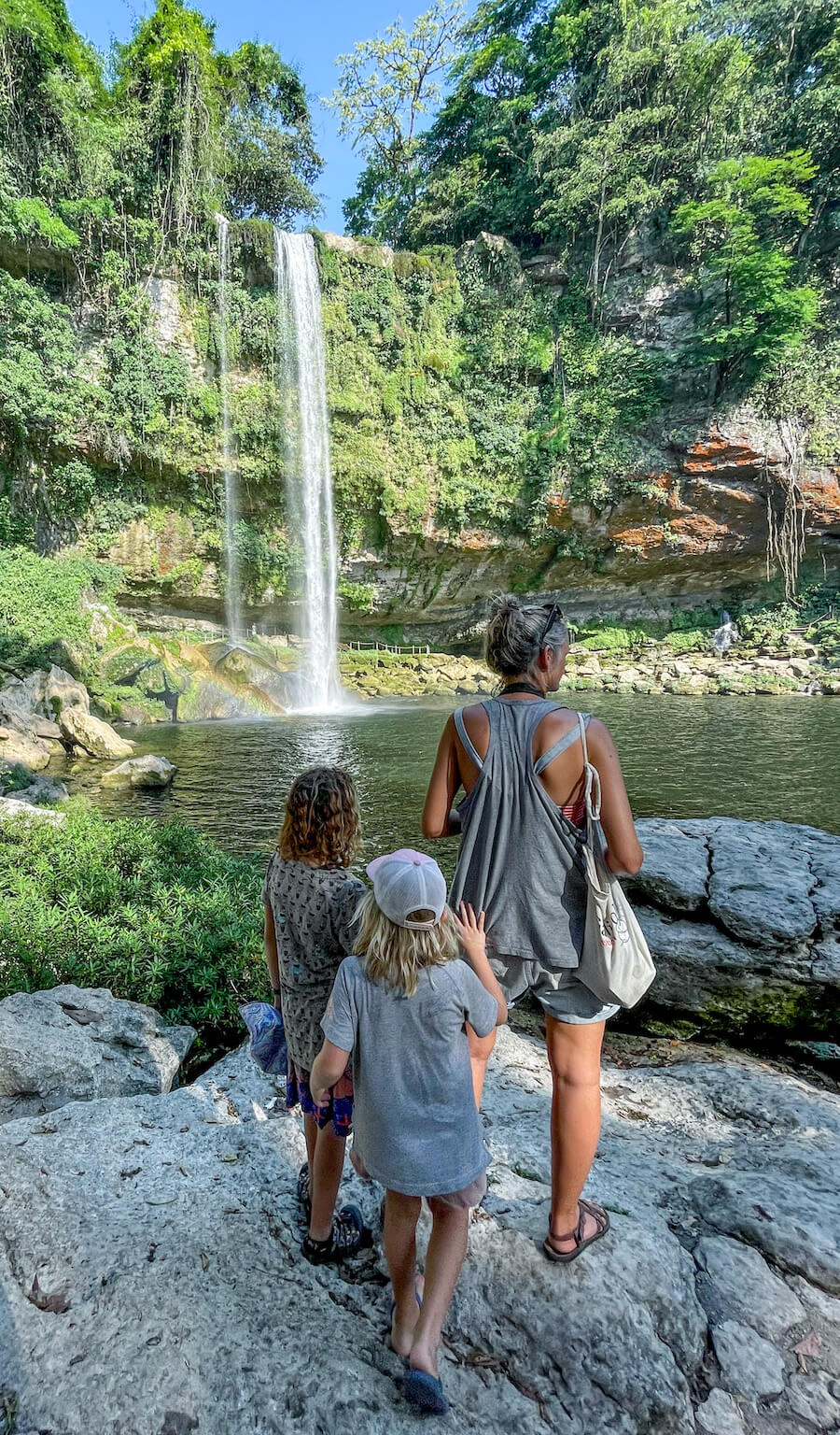 A mother and her two daughters, standing beside the lagoon at Misol-Ha, looking out at the large waterfall. 
