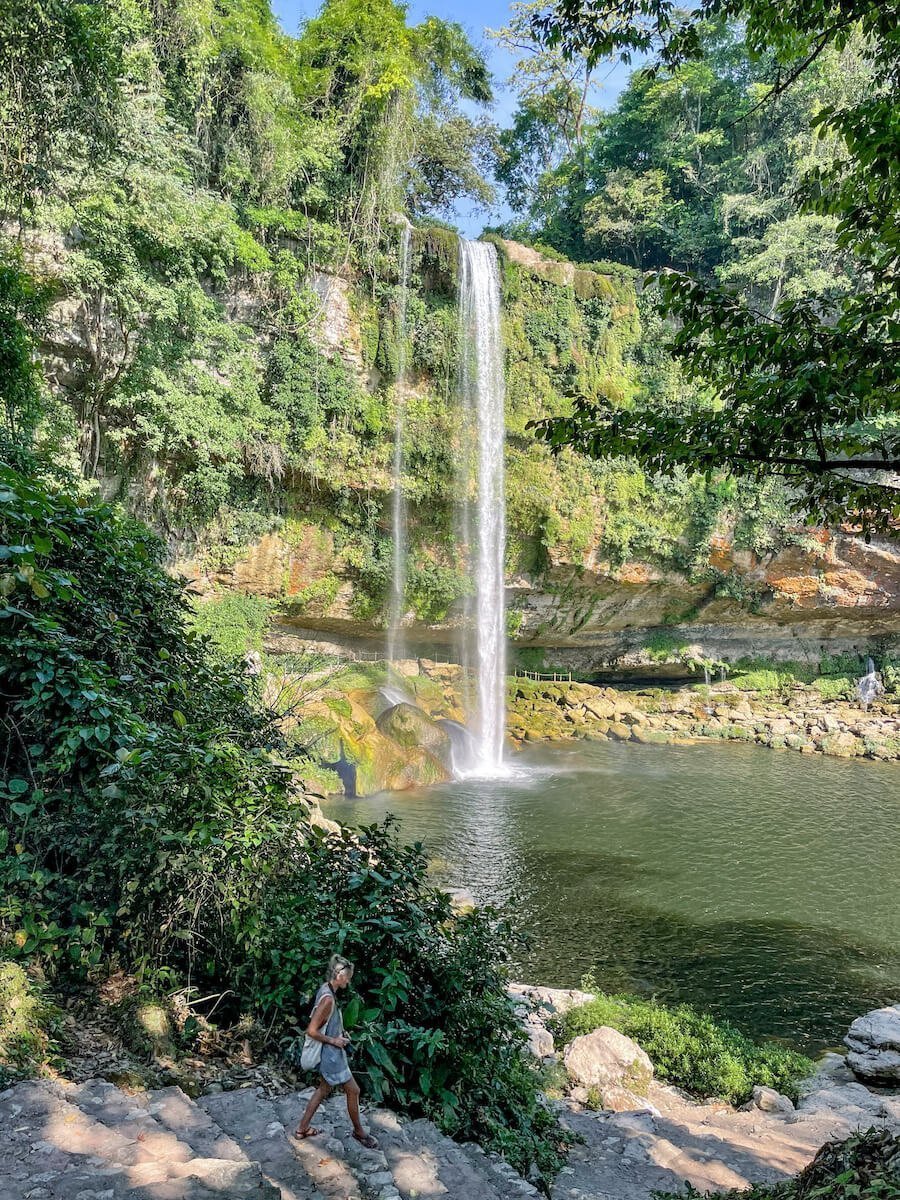 A lady walking down the steps towards the swimming hole at Misol-Ha, with the waterfall behind her.