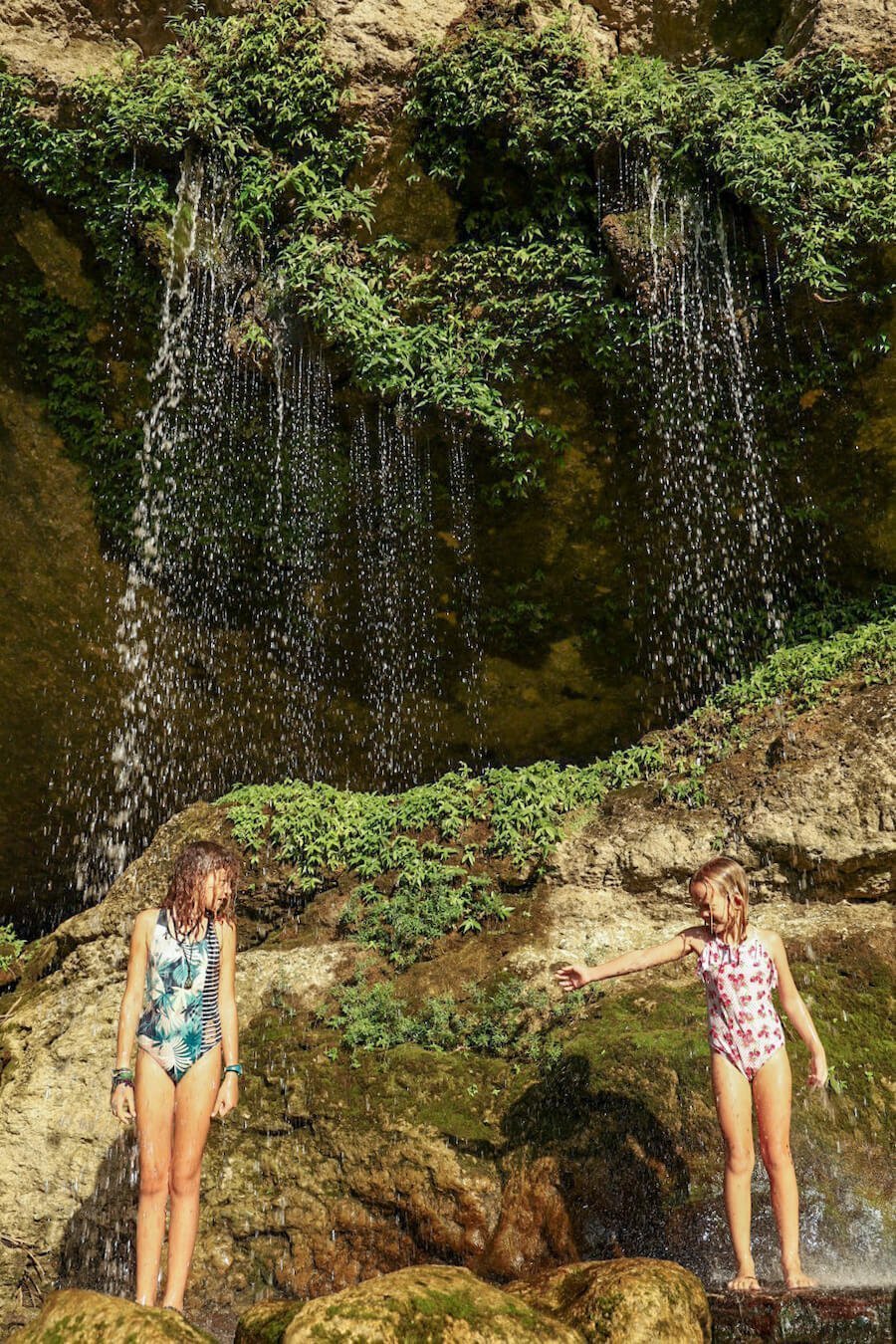 Two young girls standing under a small section of waterfall, at Misol-Ha, Palenque.