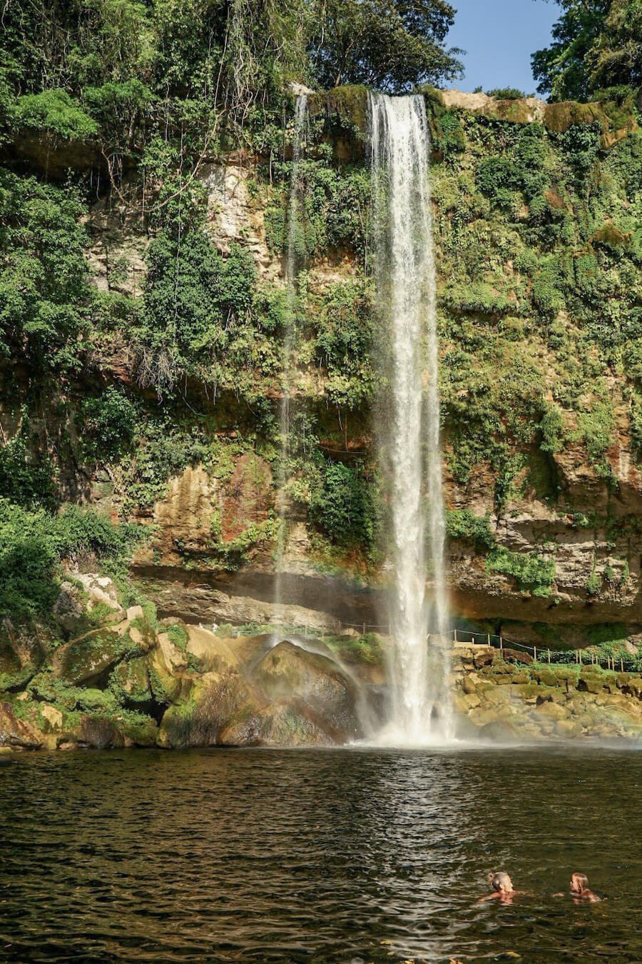 A lady and small child, swimming in the swimming hole at Misol-Ha, Palenque. With the large waterfall and small rainbow behind them.