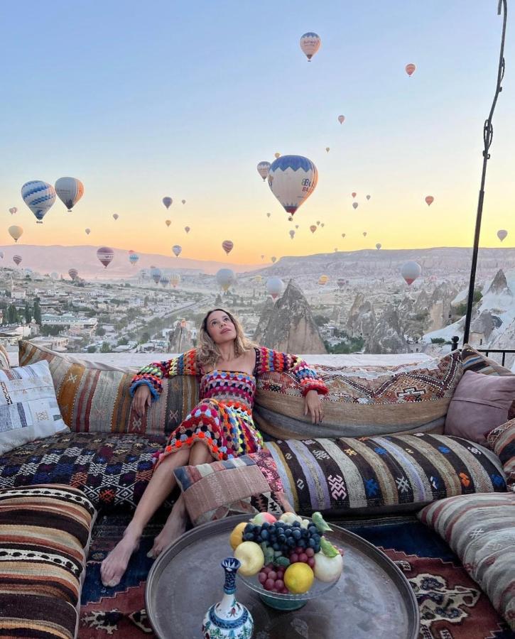 A woman relaxes on cushions on the beautifully decorated rooftop terrace at Arif Cave Hotel in Cappadocia while hot air balloons float in the distance.