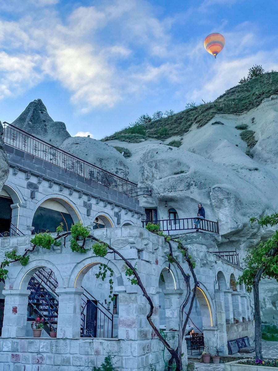 A hot air balloon flies over a cave hotel in Göreme, Cappadocia as a woman sits on the terrace enjoying the view