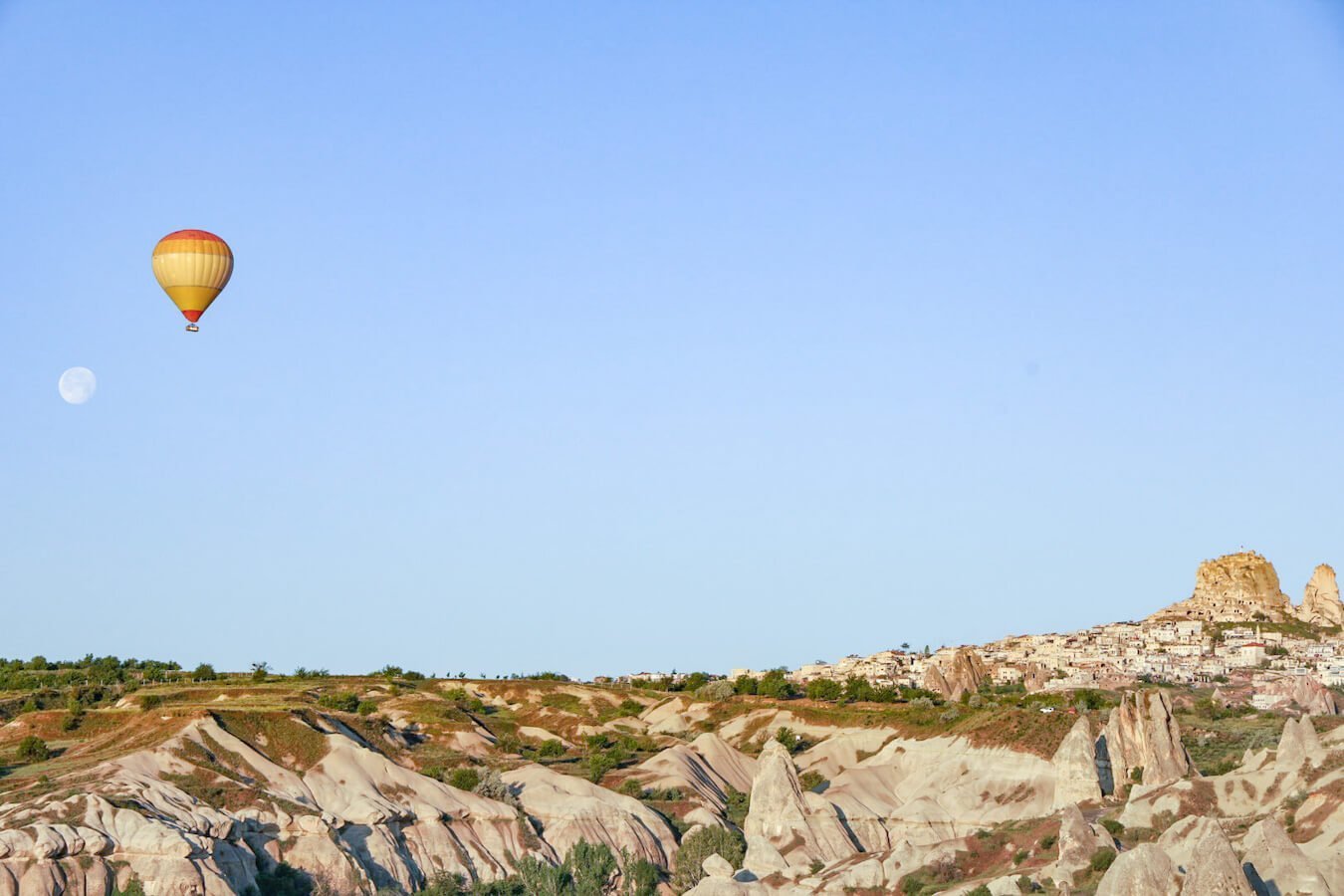A lone hot air balloon flys near Uçhisar castle in Cappadocia Goreme at sunrise. A nearly full moon is visible and incredible rock formations line the valley.