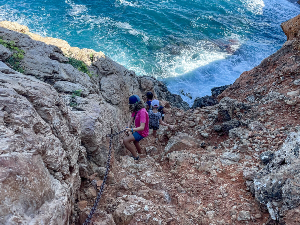 Kids scale down a rock path with a chain hand support towards the Cova Tallada sea caves