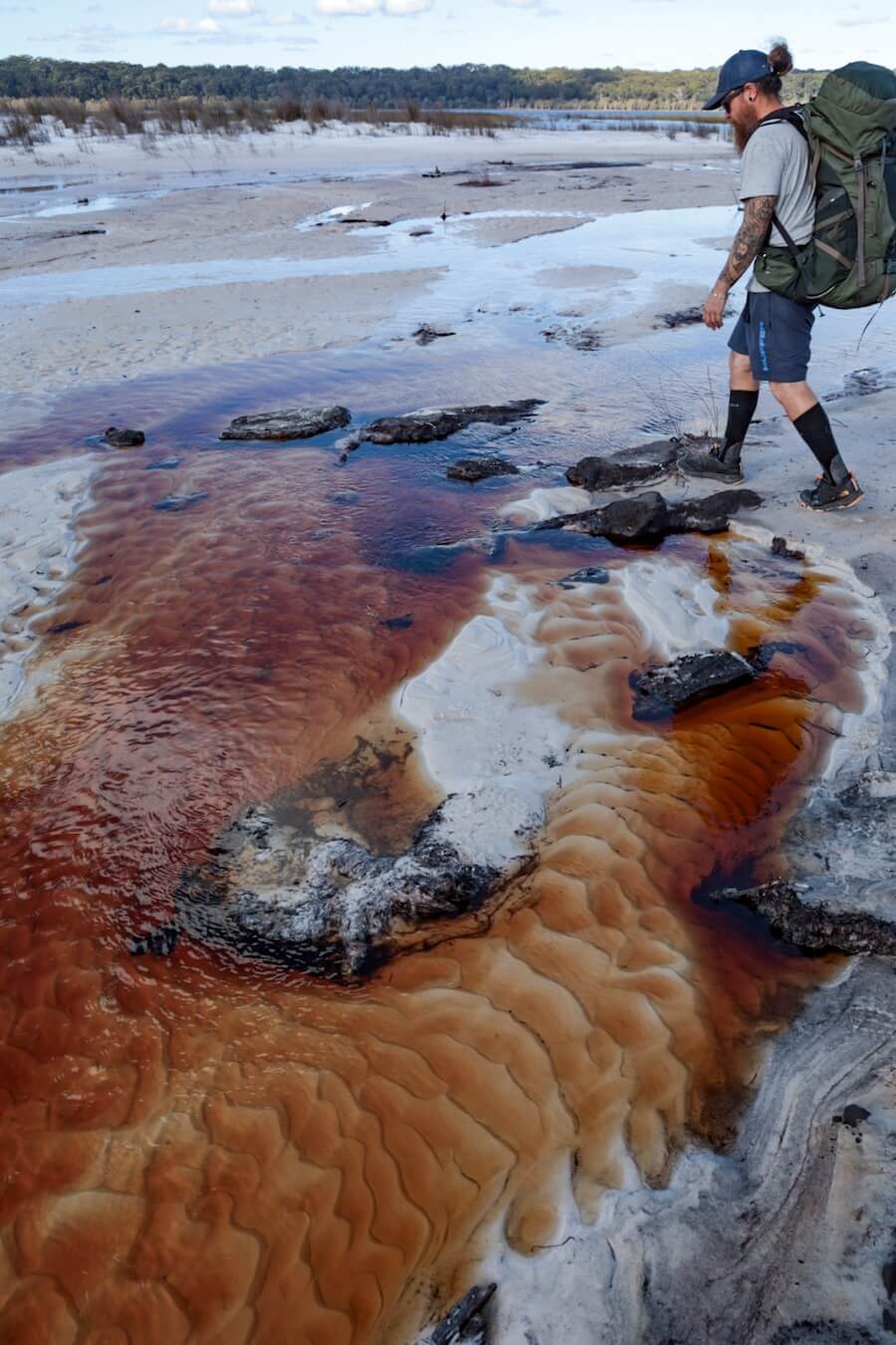 A hiker walks over a red stained stream beside Lake Boomanjin on the K'gari (Fraser island) Great Walk