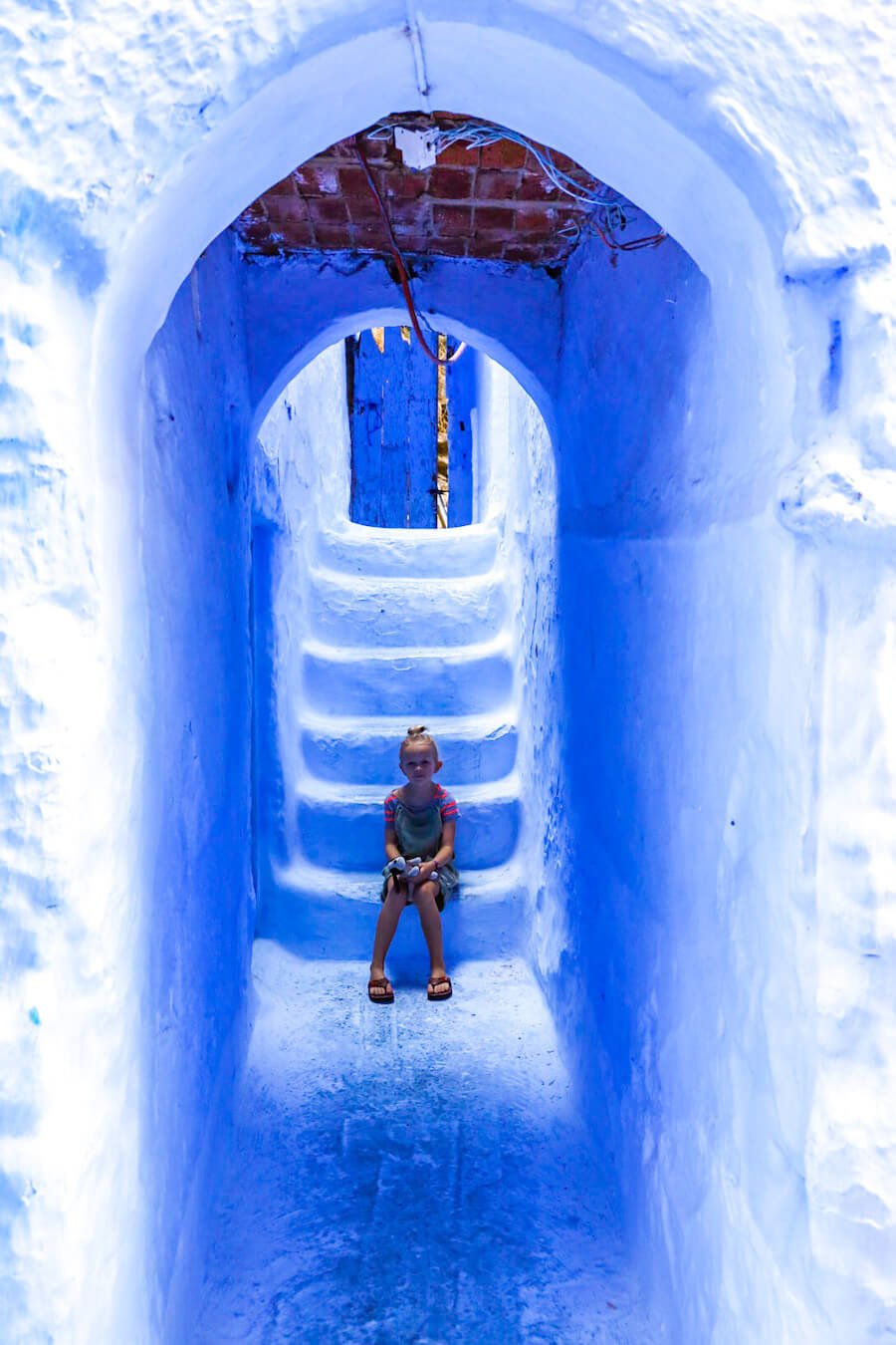 A child sits in a blue archway of the beautiful blue streets of Chefchaouen in Morocco - one of the best things to do on Morocco itinerary.