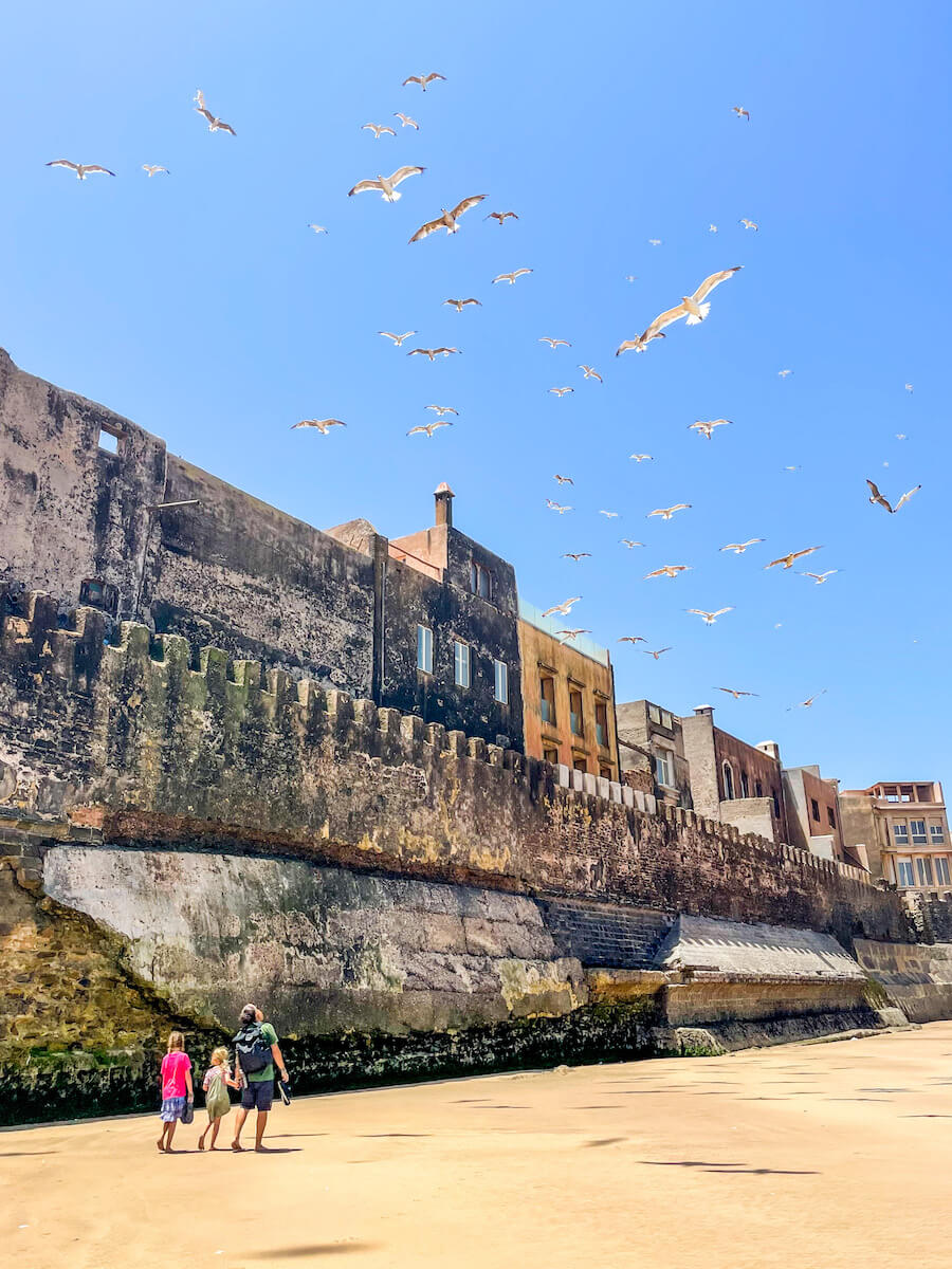 A family walk past the ramparts along the beach in Essaouira - as they travel Morocco.