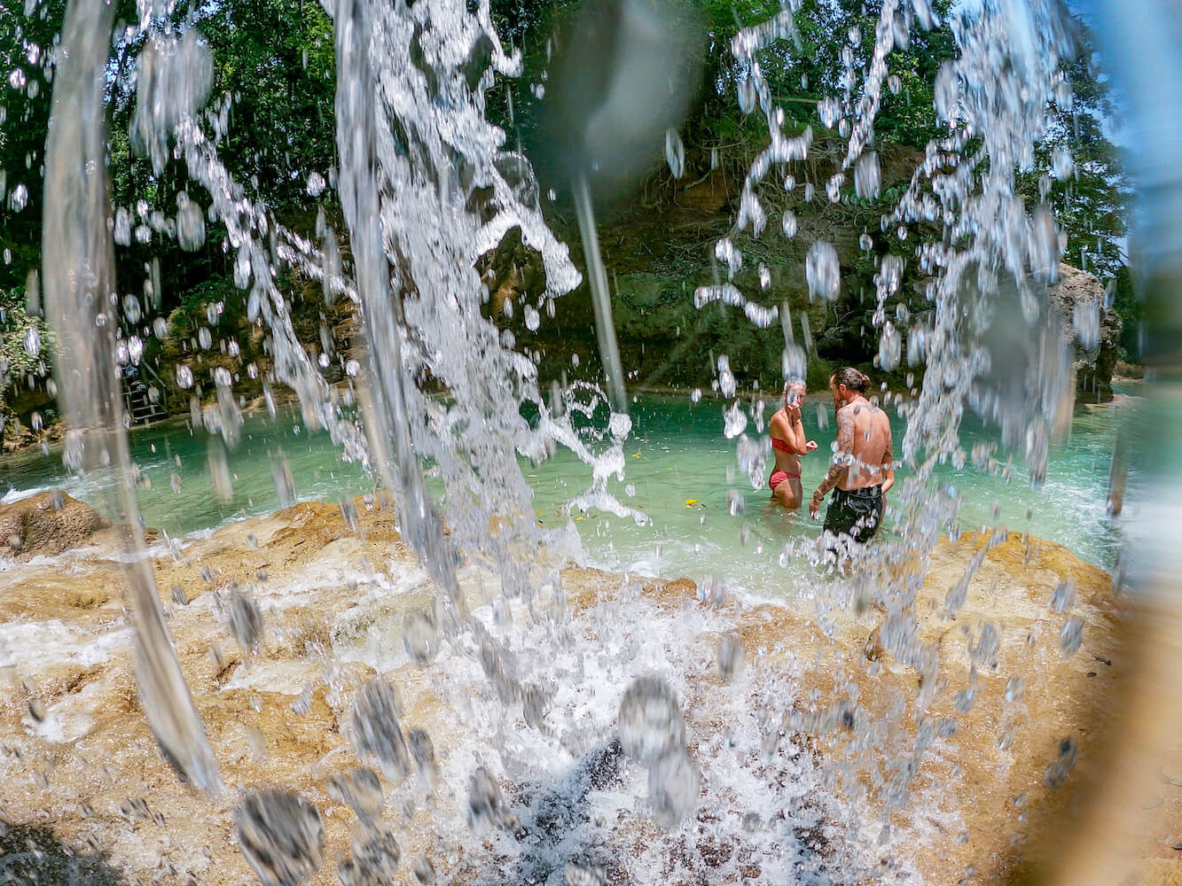 An image taken from behind a waterfall, of a woman and man standing on the edge of a natural swimming hole.