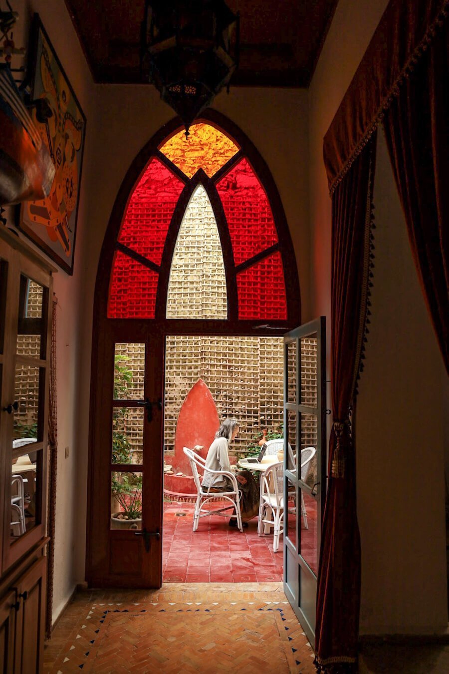 Lady works at her laptop in the outdoor courtyard of Ryad Watier.  Photo is taken through the open door and coloured glass.