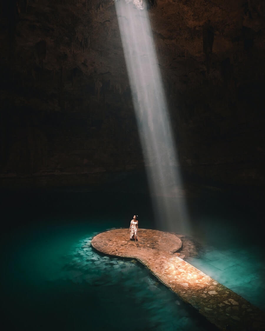 A woman stands on the platform of Instagram famous Suytun Cenote under a beam of light.