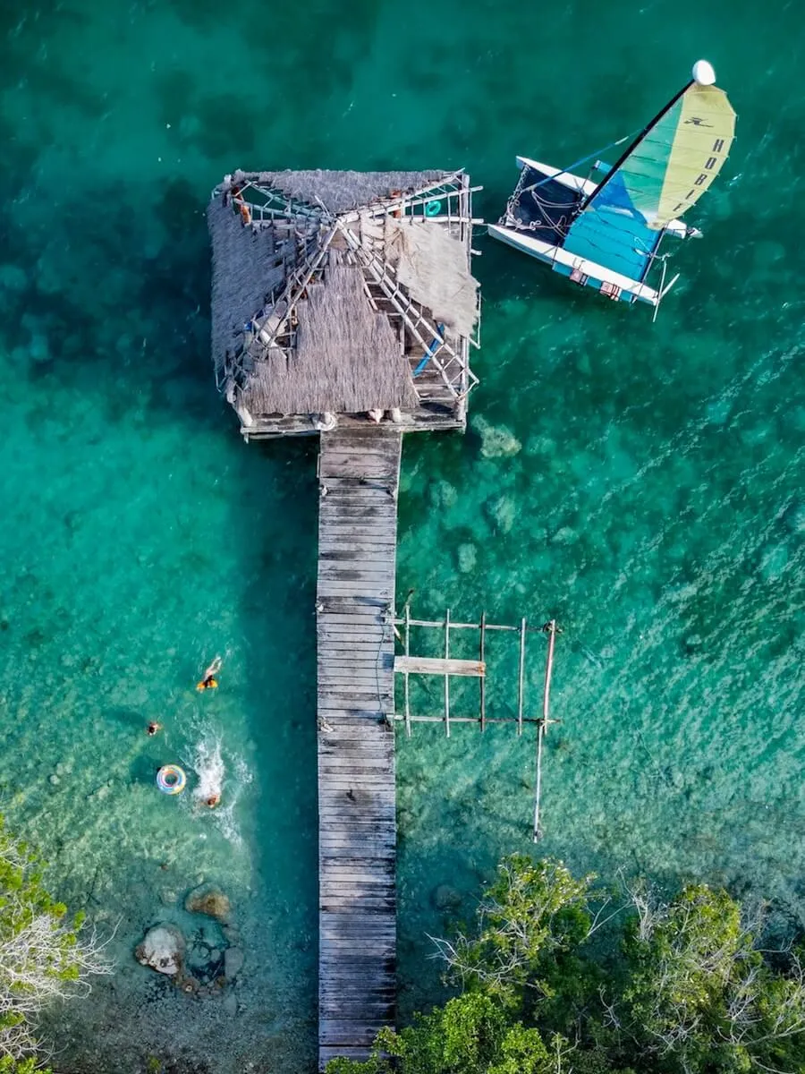 Drone image of the picturesque blue waters of Bacalar Lagoon, a palapa and pier over the water, and a sail boat, highlighting things to do in Bacalar.