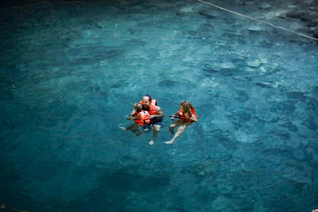 Children swim with a parent, wearing life jackets in the blue water of Cenote Samula in Valladolid