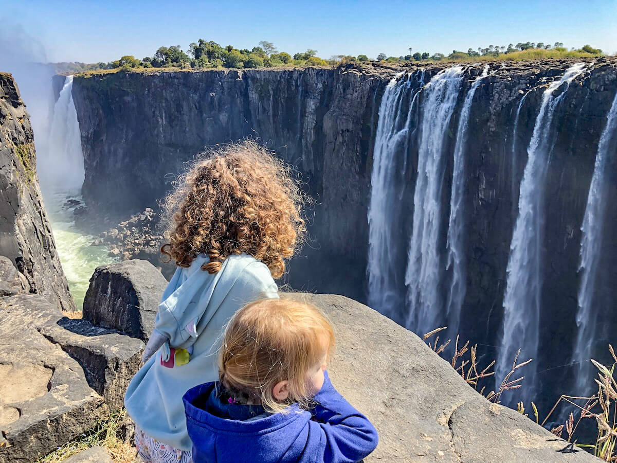 Two kids on holiday, stand beside a rock in the National Park and admire the water cascading over the Victoria Falls in Zimbabwe.