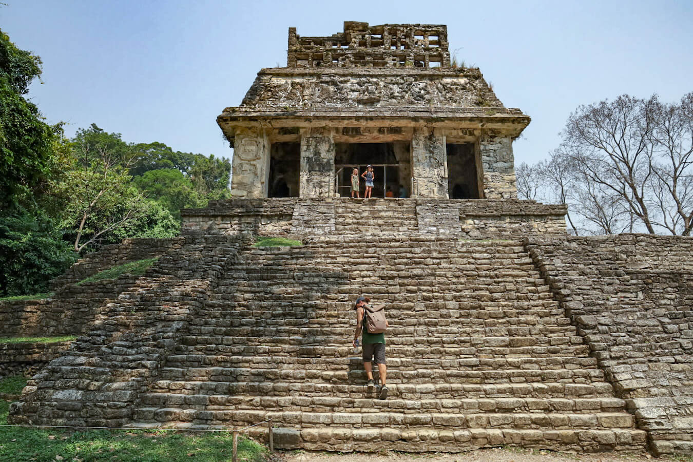 A man walking up the steps of the Temple of the Sun. Two girls standing on top in the open entrance, while on a self guided tour of Palenque.