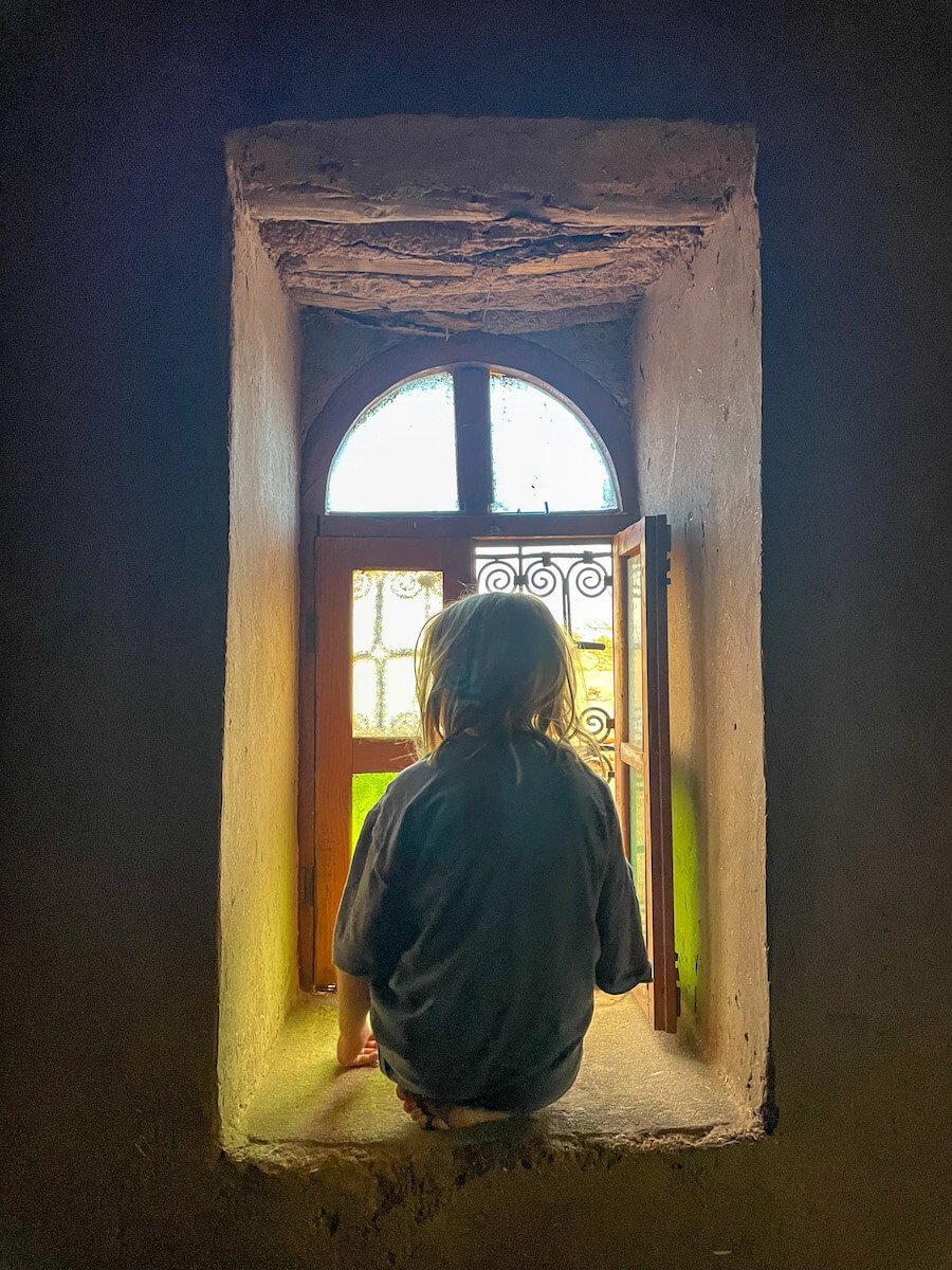 Young girl sits in the window nook of the mud brick house in Morocco, looking out to the desert.