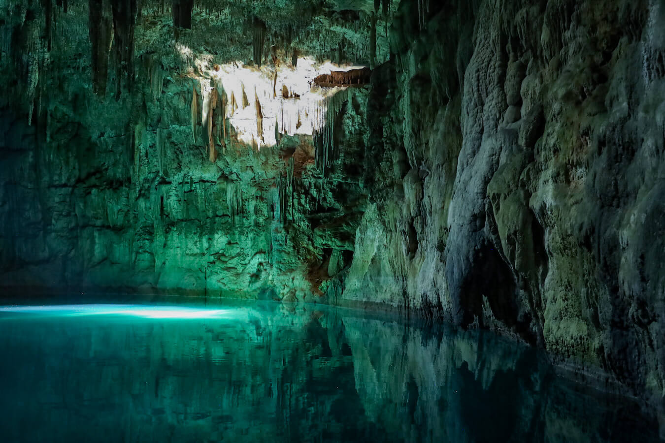 The sun beam of the closed vaulted ceiling of the Xcanahaltun cenote lets in enough light to turn the cenote walls and water a bright crystal blue.