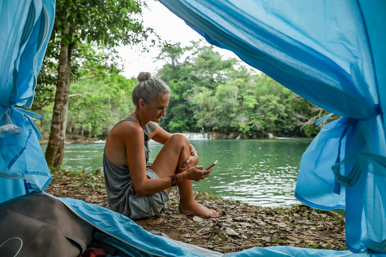 Photo taken from inside a tent, looking out at a lady sitting beside a river, with a small waterfall behind her, in the Lacandon Jungle.